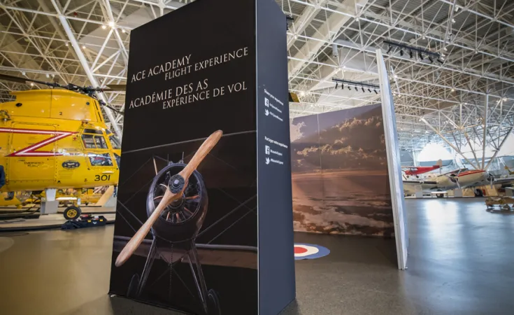 Un grand panneau d’exposition avec le titre « Académie des as, expérience de vol » et une image d’un biplan. Derrière le panneau, à gauche, se trouve un hélicoptère de sauvetage jaune.