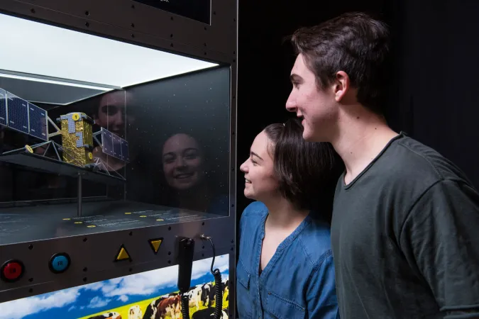 Deux personnes observent attentivement la maquette d’un satellite présenté dans la vitrine d’un module d’exposition.