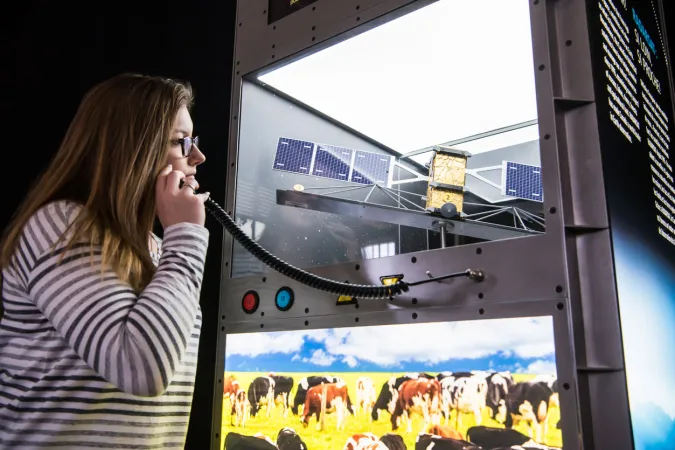 Une femme tient un dispositif d’écoute contre son oreille. Le module d’exposition devant elle contient la maquette d’un satellite et une photographie rétroéclairée de vaches dans un champ.