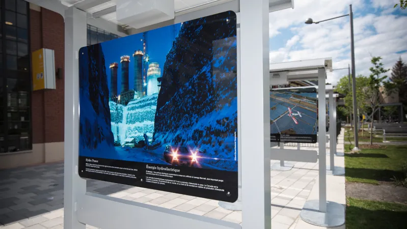 Cascading panels of a photographic exhibition is set up outdoors. The first photograph is of a hydro power plant in British Columbia, during the winter. There is white text below the photograph.