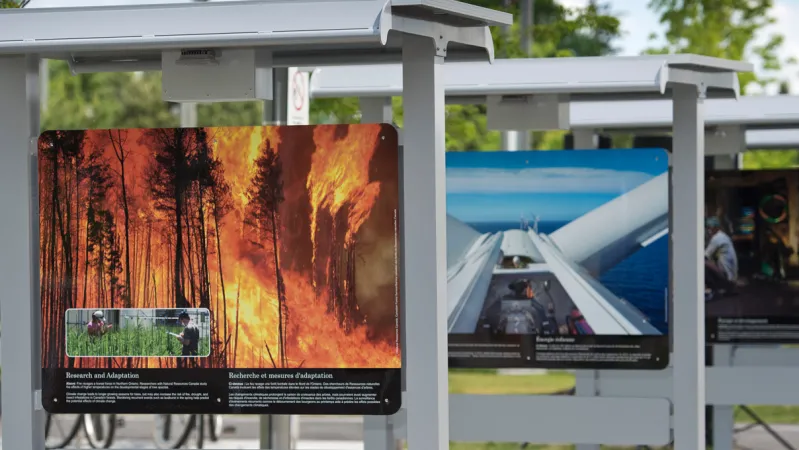 A panel from a photographic exhibition that shows a forest fire. There is a smaller inset photograph that shows two people standing in tall grass. There is white text below the photograph. The second panel is a photograph taken from the top of a wind turbine.