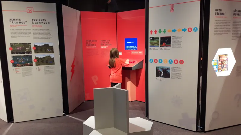 An exhibition module with gray and orange panels with colour graphic icons, texts and images can be seen in the background. A child with an orange shirt is playing a video game. A gray seating structure can be seen in the foreground.