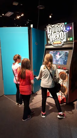 Trois enfants jouent au jeu d’arcade Guitar Hero. Un panneau bleu se trouve à leur gauche.