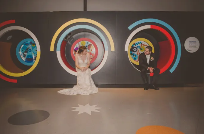 A cute picture of a bride and groom sitting in the kids’ section of the Canada Science and Technology Museum. They are sitting in two circular seats outlined in bright red, white, blue, and yellow colours on a black background and looking at each other.