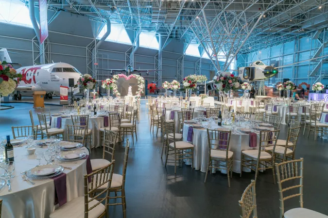 An indoor view of tables with white table cloths with violet accents and a white airplane in the background. Large tables are set up around the room with elegant white chairs. The décor is very light green and pink floral and there is a heart-shaped flower arch at the front of the room.