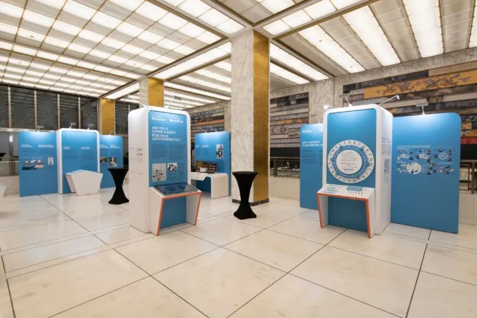 De nombreux modules d’exposition sont installés dans une grande salle comportant trois grosses colonnes. Les modules sont bleus et blancs.