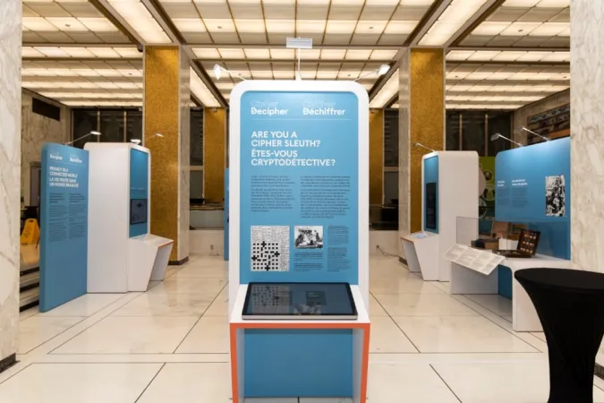 Many exhibition modules are on display in a large room that has large white columns. The modules are blue and white. The module in the center of the room has the title Are you a cipher sleuth. Below the text is a screen lying flat on a table that is attached to the panel.