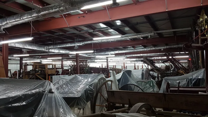 A series of carriages and farm equipment sit in a cramped warehouse.