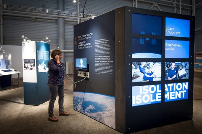 A museum visitor holds an earcup to their ear as they stand beside a large exhibition module. The module contains a wall of TV screens in addition to the listening station.