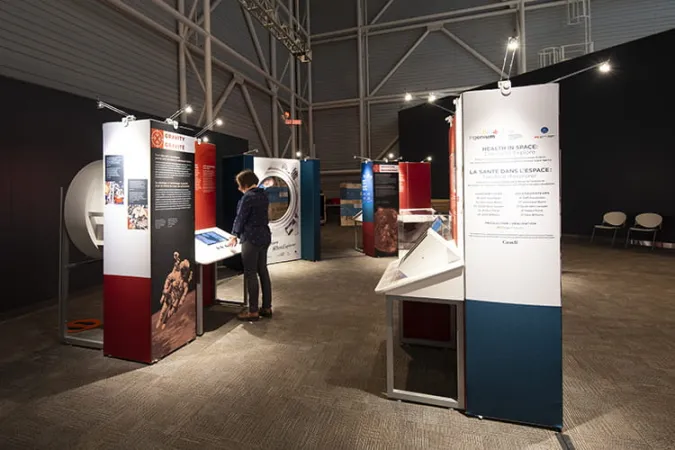 Quatre modules d’exposition aménagés dans une grande salle. Une personne se tient devant une exposition interactive.