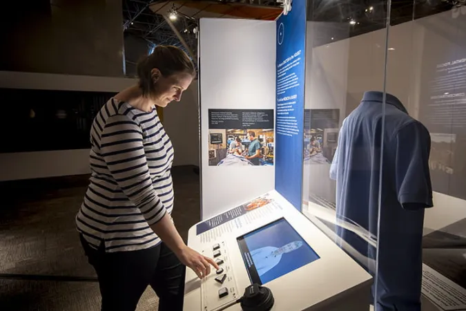 A museum visitor leans in to press buttons on an exhibition interactive. A person is visible on the TV screen.