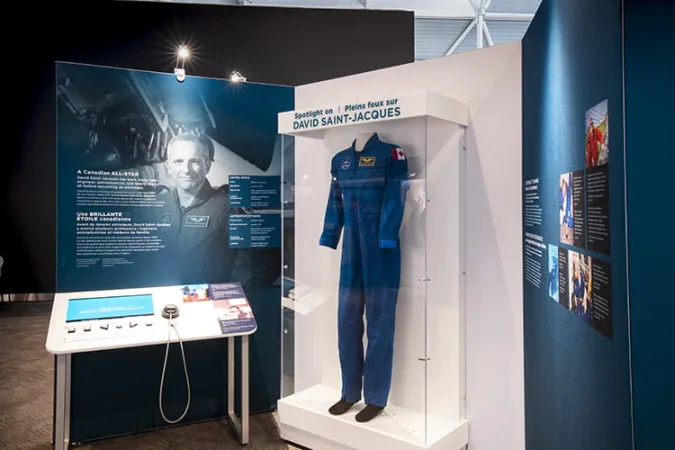 A curving exhibition module shows at its centre a blue flight suit within a display case. Overtop the suit, there is the heading “Spotlight on David Saint-Jacques”.
