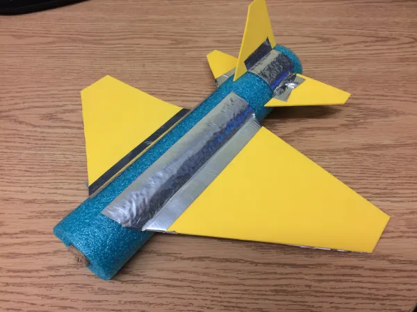 A pool noodle airplane with wings and a tail made out of yellow foam and pieces of cardboard.