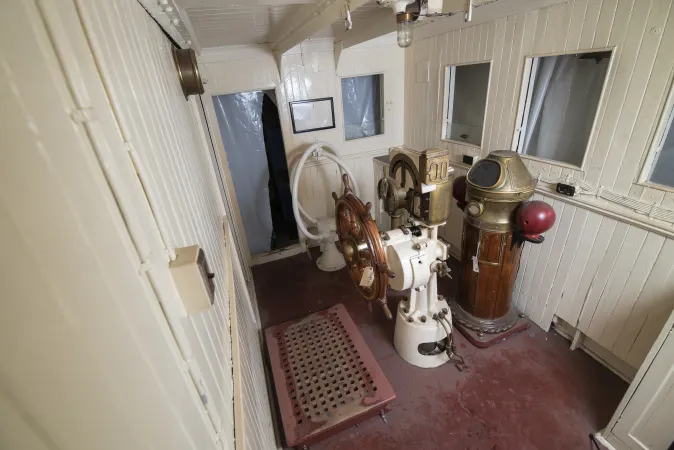 An aerial view of the steering wheel and other pieces of equipment, inside the wood-panelled walls of the pilot house.