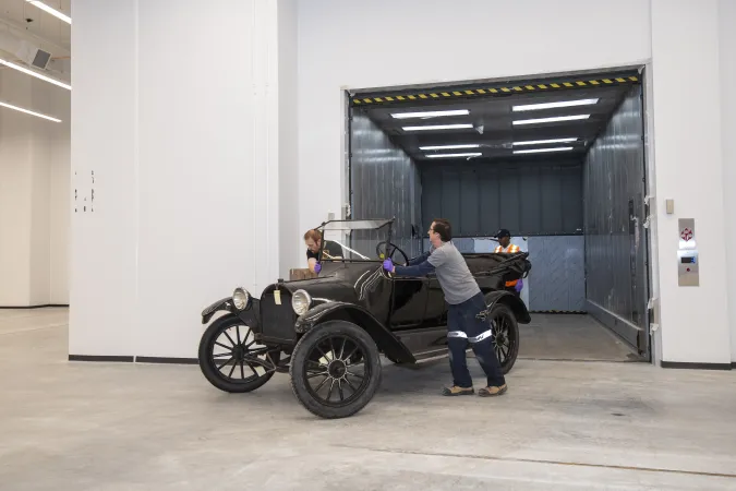 Two men wearing artifact gloves move a vintage black car off an oversized elevator.