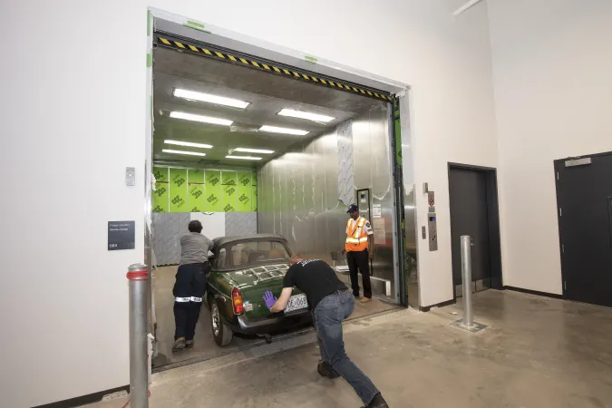 Two men push a green sports car into the oversized freight elevator.