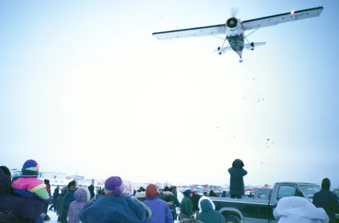 Un groupe de personnes se tenant debout à l’extérieur observent un petit avion blanc larguer des bonbons et des cadeaux tandis qu’il survole la foule.