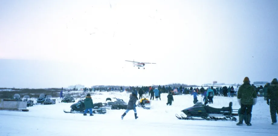 A group of people stand outdoors, looking up as a small white plane drops candy and gifts while it flies over the crowd.