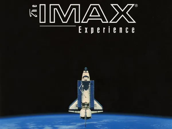 A white and blue space shuttle leaves the Earth’s atmosphere and is pointing upwards to the words “The IMAX Experience.”  The image background is black, the text is white, and we can see a blue ocean with some white clouds in the Earth at the bottom of the picture.
