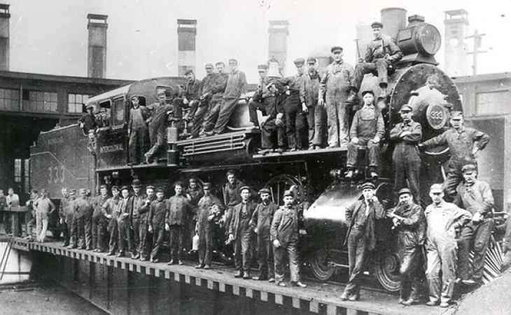 Image is a black-and-white photograph showing employees standing on and around a steam locomotive. There are about 40 men