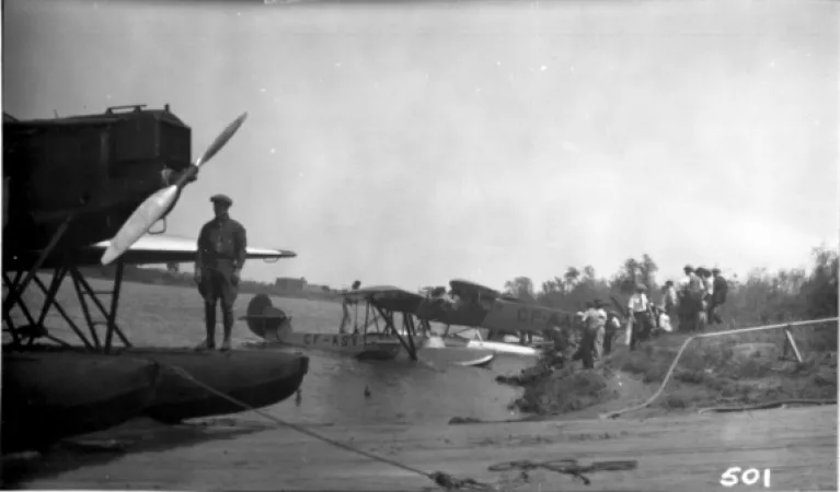Image is a black-and-white photograph showing three sea planes at a shore where people are standing. In the foreground, one man is standing on one of the floats of the closest plane.