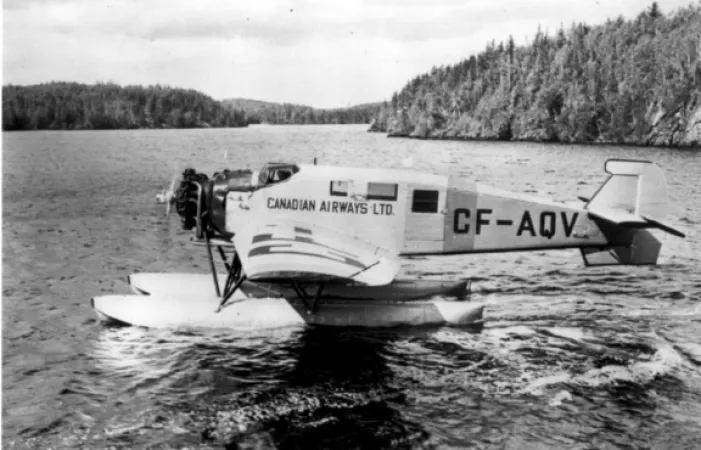 Image is a black-and-white photograph showing a seaplane in a lake with shore or islands covered in trees in the background. “Canadian Airways Ltd” and registration number “CF-AQV” shows on the side of the aircraft.   