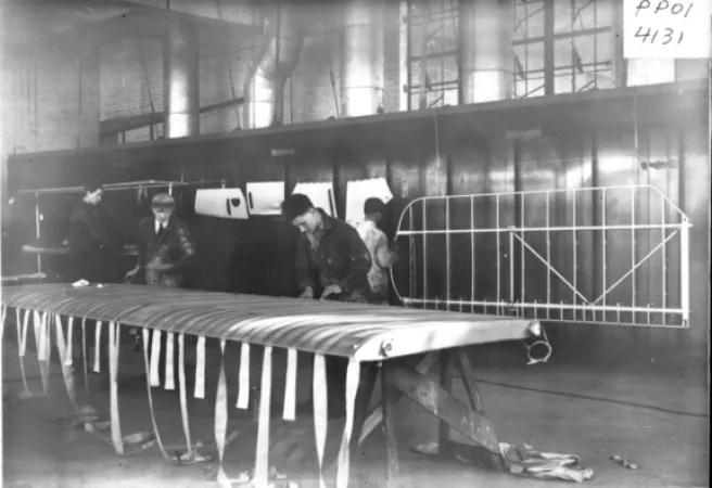 Image is a black-and-white photograph showing a shop floor, where men are building aircraft wings. Two men stand behind the frame of a wing propped up horizontally with strips of fabric visibly hanging from it. 