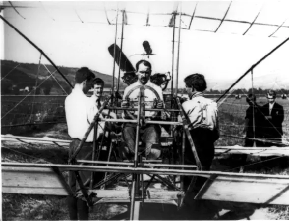 Photographie en noir et blanc montrant un homme assis aux commandes d'un premier avion avec un cadre ouvert. Ses collègues de l'Aerial Experiment Association se tiennent autour de lui.