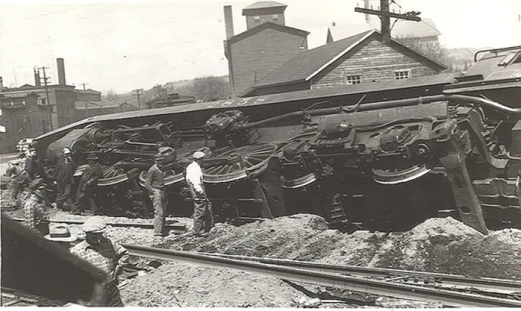 Image is a black-and-white photograph showing a steam locomotive that has derailed and is lying on its side. Men in the foreground stand looking at the wreck.