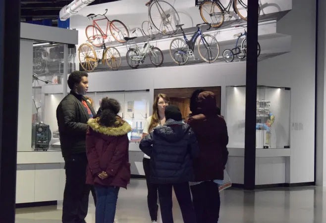 A group of teenagers and one coordinator gather in a circle to sort out their ideas and problem solve. Behind them is a showcase of the different kinds of bikes there have been through time. 