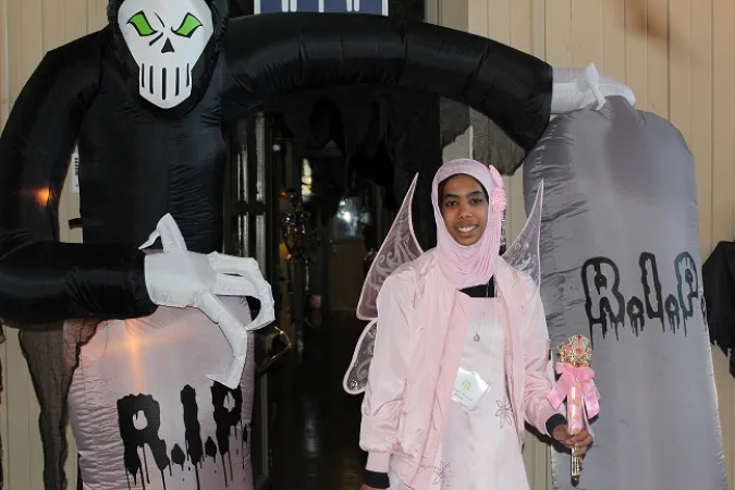 Une jeune fille vêtue de rose et d’un hijab assorti porte des ailes de papillon et pose devant un décor d’Halloween.