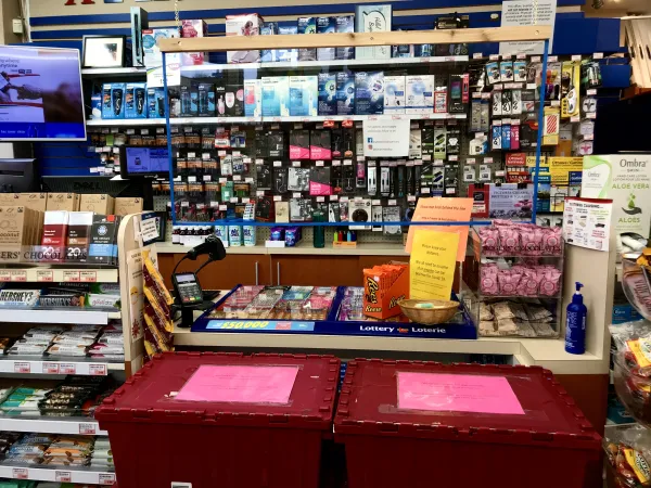 A checkout counter at a pharmacy; several red plastic boxes with a pink notice indicate that customers should not move the barrier in order to protect the safety of the staff, and a plexiglass sheet is suspended to separate customers and check out staff.