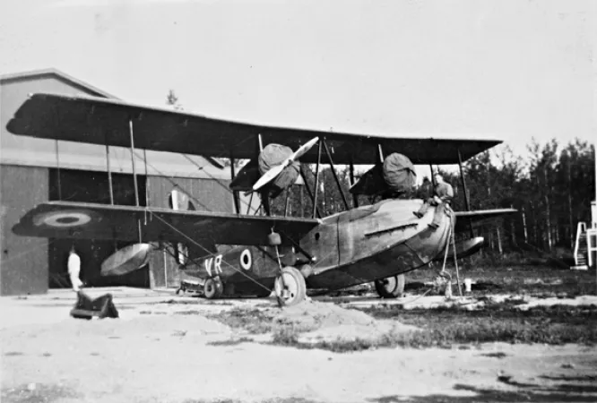 Image is a black-and-white photograph showing a twin-engine Vickers Vancouver II bi-plane in front of a hanger. A man is sitting on the nose of the aircraft.