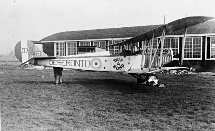Image is a black-and-white photograph showing a side view of a Curtiss JN-4 aircraft in an airfield. “Deseronto” and “buy a bond” are written on the side of the plane. 