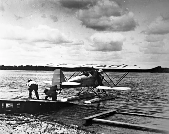Image is a black-and-white photograph showing a seaplane at the end of a dock. Two men are on the deck and are holding on to the aircraft with a rope. 