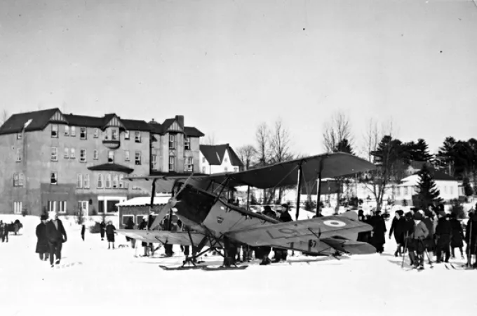 Image is a black-and-white photograph of a biplane aircraft in a snowy field in front of a large hotel. People surround the aircraft, some of them are on skis. The aircraft is a de Havilland D.H.60 Moth and also has skis. 