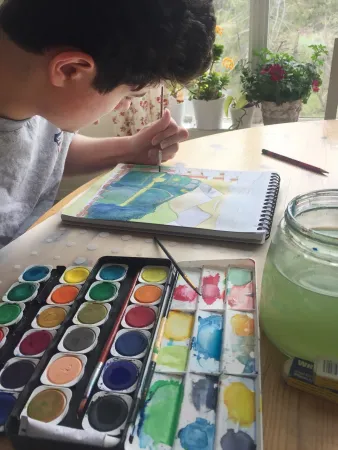 A boy leans over his art pad, intently painting a colourful picture. A paint palette is visible in the foreground, and some flowering plants and a window can be seen in the background.