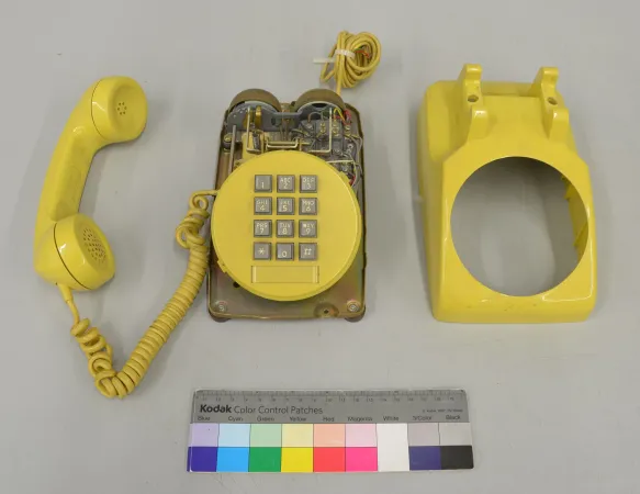 A yellow, lustrous plastic desk phone with matte grey keys, cover removed exposing wiring, sits on a grey background. 