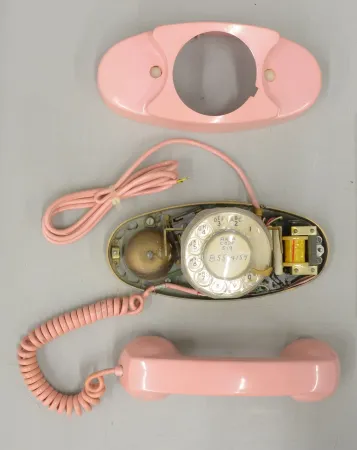 A pink rotary phone with the plastic case removed, exposing the dial and bell, sits on a grey background. 