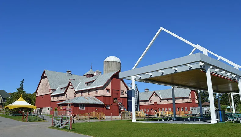 A wide shot of two large, red-and-white agricultural buildings and a navy blue silo. Green grass and a covered, outdoor picnic area are visible in the foreground, and a blue sky in the background.