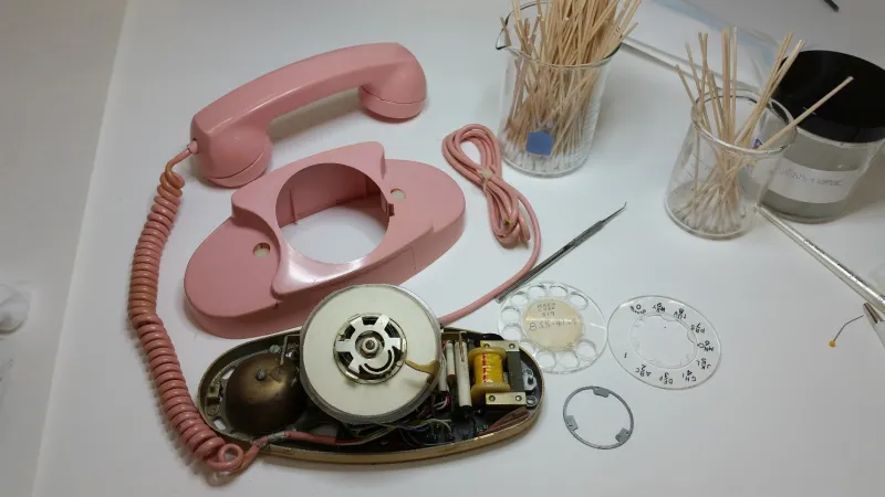 A pink rotary phone, partially disassembled, sits on a work bench with a variety of tools. 