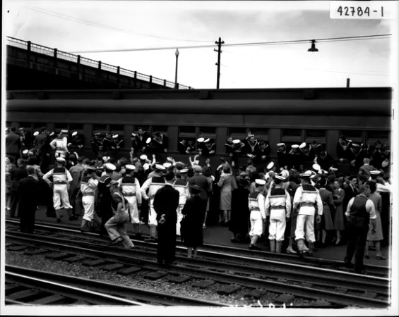 L’image est une photo noir et blanc montrant une voiture de train et une foule de soldats en uniforme de la marine qui attend à côté de la voie ferrée, faisant ses adieux aux compagnons déjà montés à bord et qui regardent par les fenêtres. 