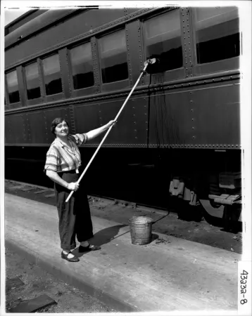 Image is a black-and-white photograph of a woman cleaning the outside of a train car with a long mop. 