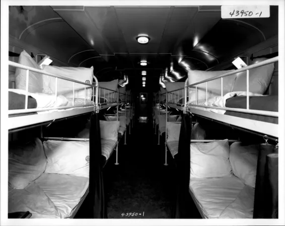 L’image est une photo noir et blanc montrant des rangées de lits superposés alignés de chaque côté d’un nouveau wagon. 