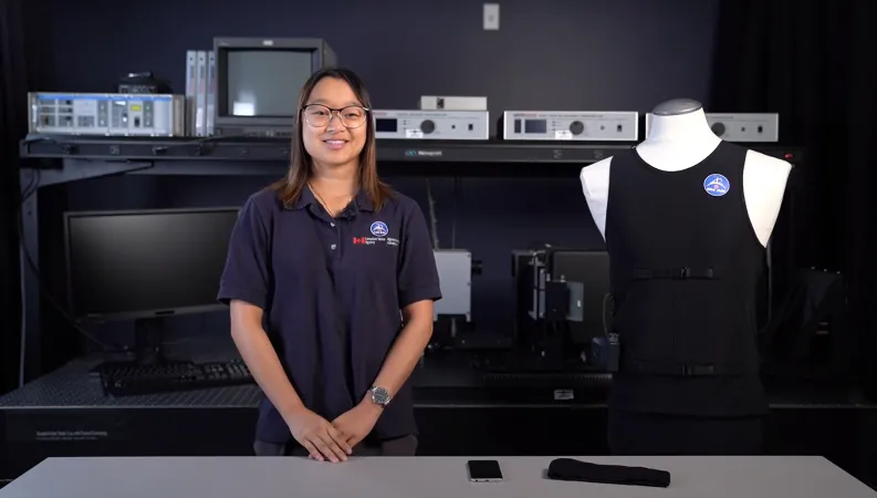 A young woman wearing a navy blue shirt smiles as she stands next to a mannequin dressed in a black tank top. A computer and a variety of equipment is visible on shelves in the background.