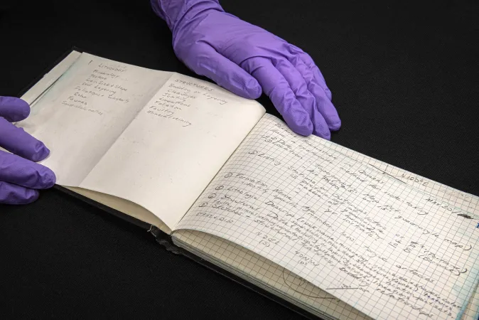 A rectangular book with writing sits on a dark surface, held open by two purple gloves. 