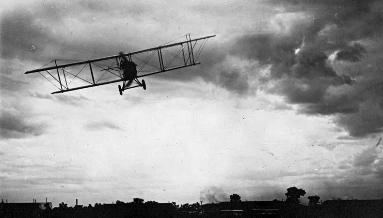 A black-and-white image depicts a front view of a biplane flying over flat land.