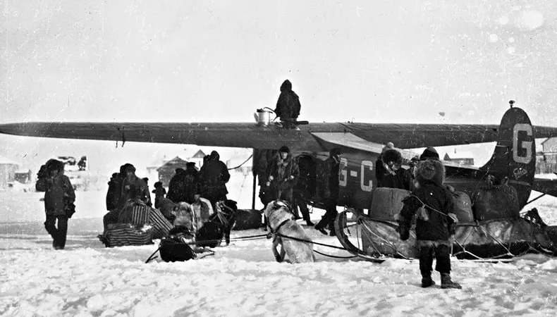 Une image en noir et blanc montre un avion en train d'être déchargé. Il y a plusieurs personnes qui le déchargent, avec un traîneau à chiens qui attend.