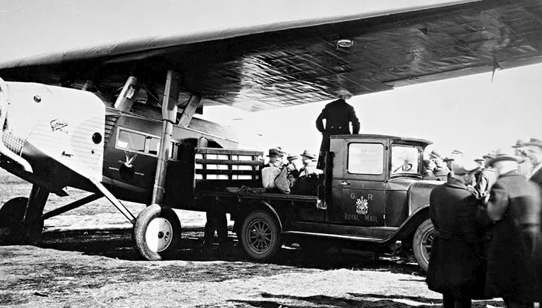 A black-and-white image shows a dozen men standing around a truck and an airplane.
