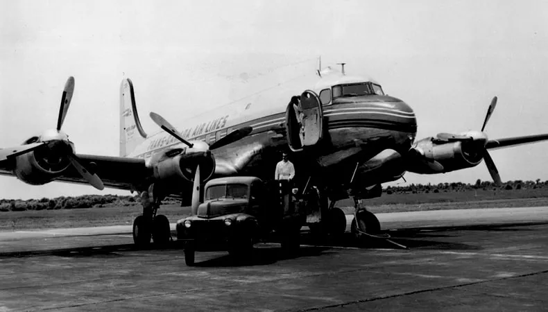 Une image en noir et blanc montre un gros avion avec sa porte ouverte, ainsi qu’un camion et un homme. 